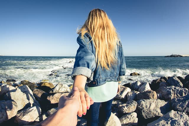 岩の転がる海辺を男性の手を引いて歩く金髪の女性の#followmeto画像